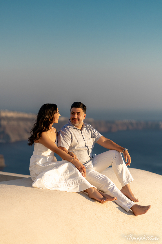 A couple shares a serene moment during a photoshoot in Santorini, enjoying the beauty of the sunset as they sit together on a rooftop with the island's cliffs and sea in the background.